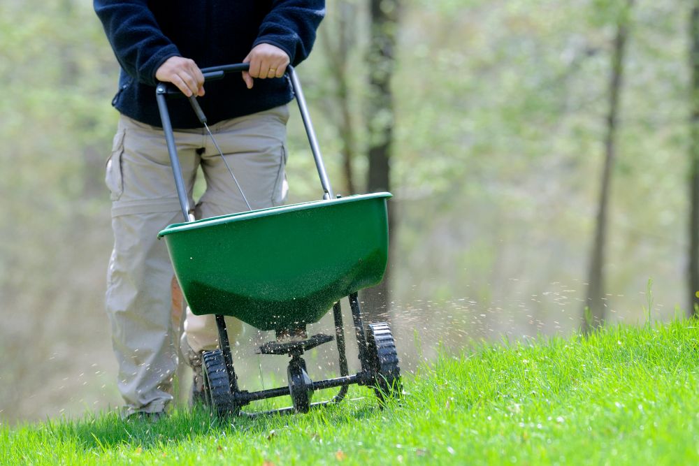 A man uses a wheelbarrow to spread grass, with a van parked in the background.