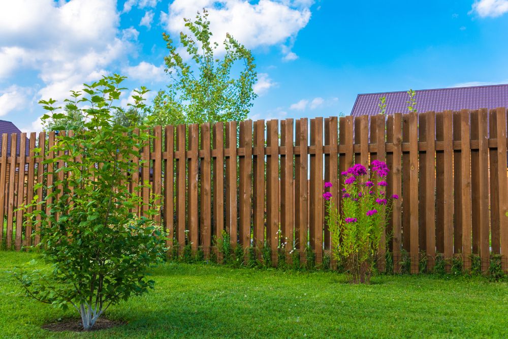 A wooden fence surrounds a yard featuring a tree and colorful flowers blooming nearby.